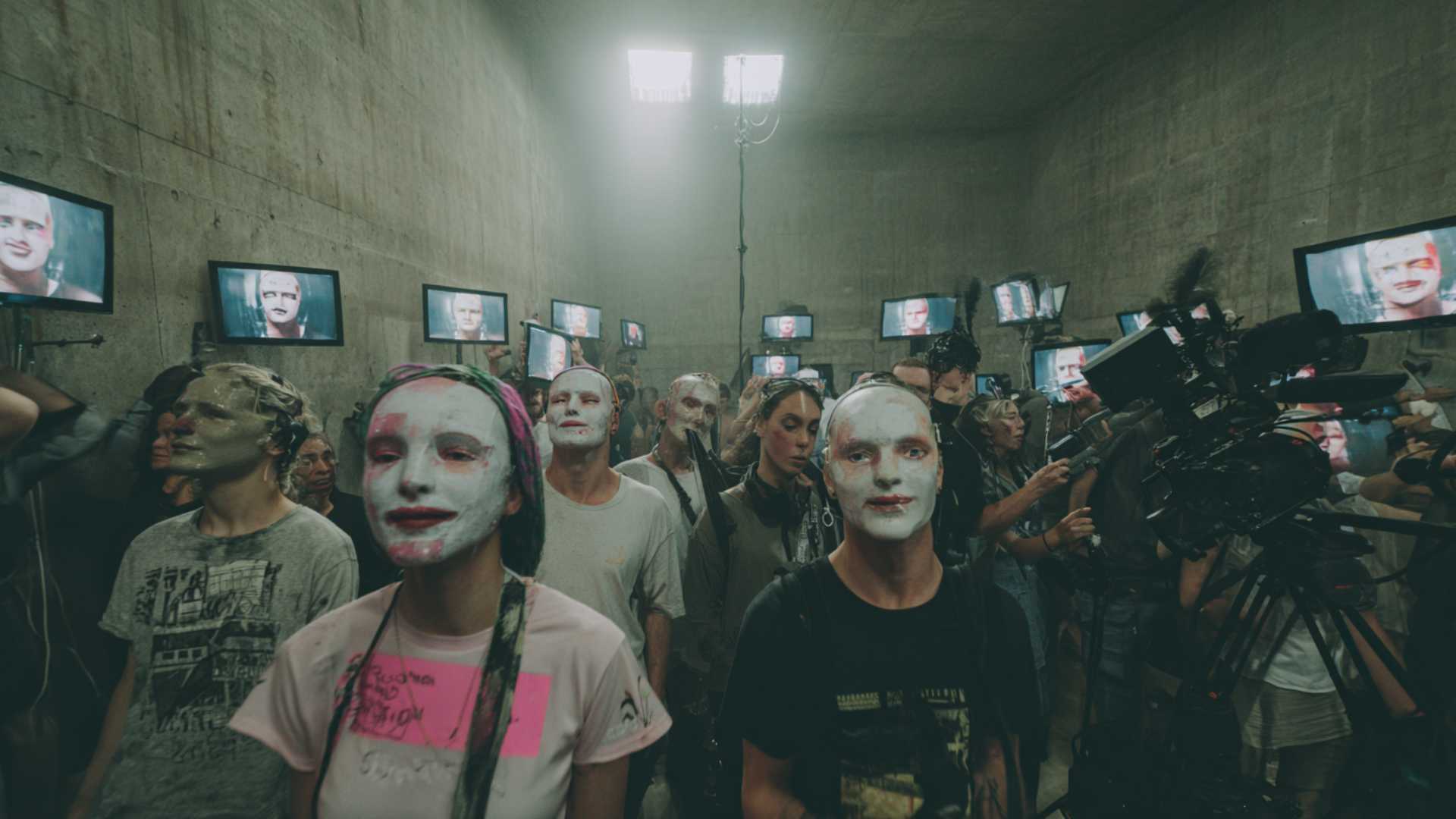 Rows of motionless figures face a wall of glowing video screens in a concrete room.