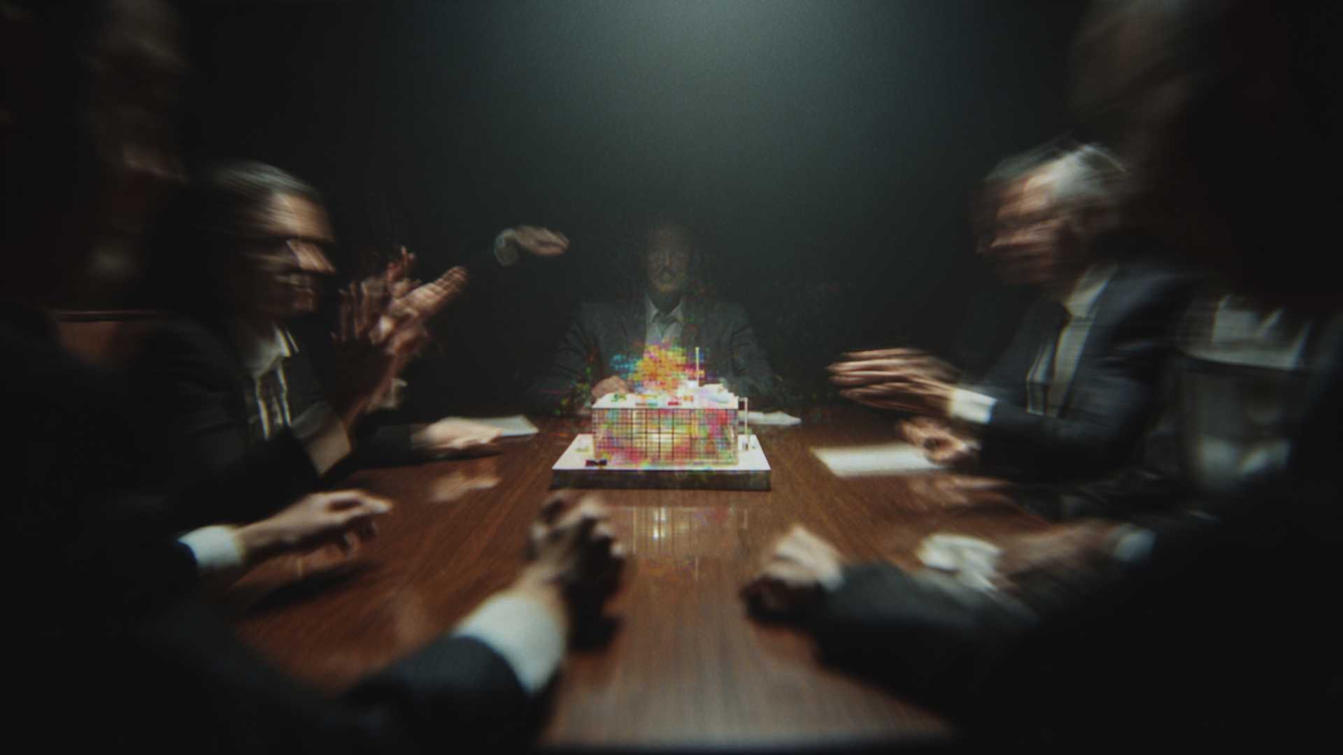Blurred figures sit around a long table, facing a brightly lit model at its centre.